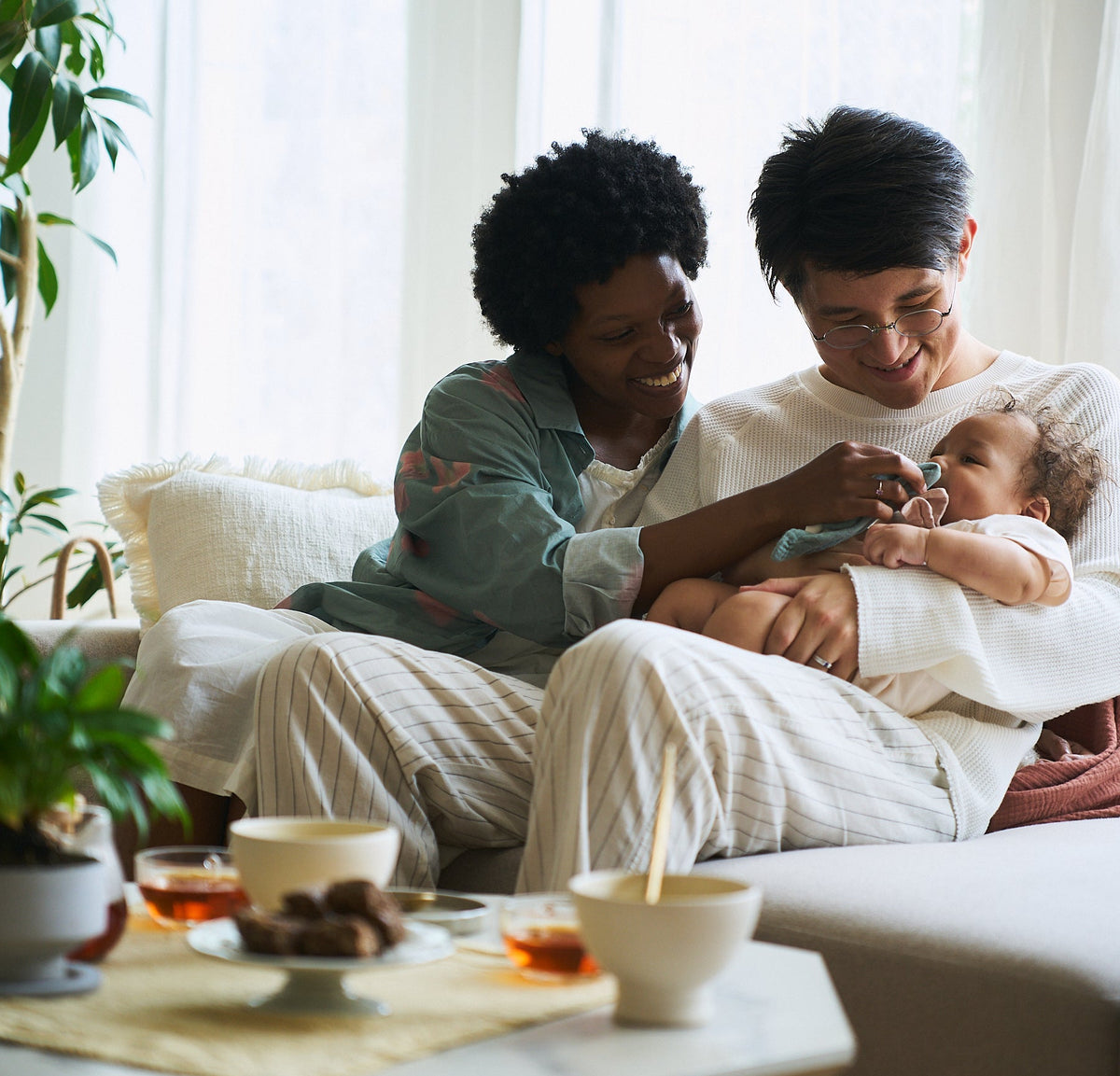 A man and a woman sitting on a sofa in a bright living room, with the man holding a baby. A coffee table with cups and a plant is visible in the foreground.